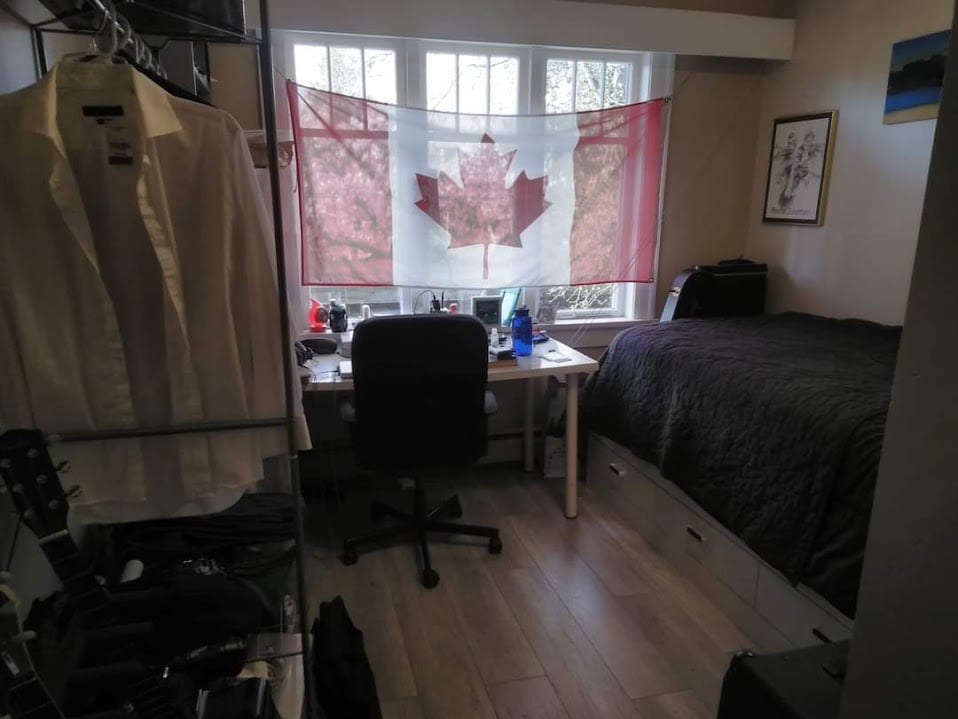 A bedroom with a desk at the window, a dark bedspread, and guitars stored along the left.