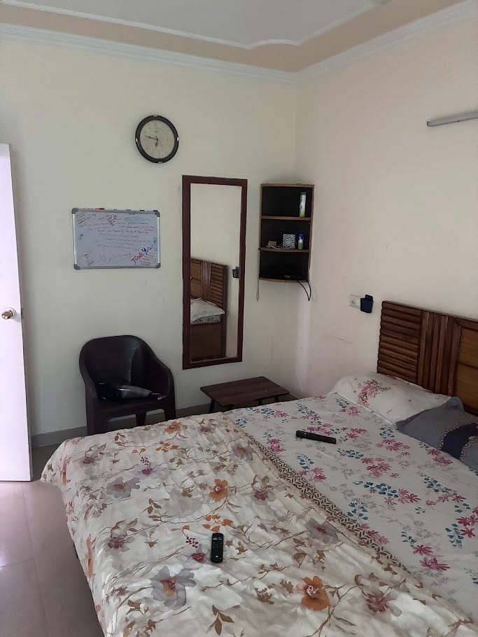 The same floral bedroom from the other side, with a mirror, a chair and a small wall shelf.