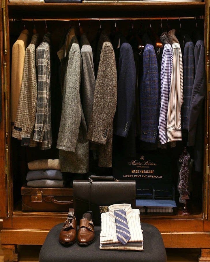 An open wooden wardrobe with jackets and shirts hung in groups, shoes and a folded shirt in front.