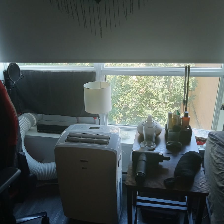 A window with a portable air cooler, a lamp, and a side table of everyday objects.
